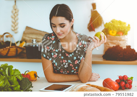 An attractive young dark-haired woman holding green apple and choosing the recipe for a delicious meal while smiling in sunny kitchen. Tablet pc is the best cookbook 79551982