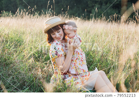 Mother, sitting on the grass in the field outdoor and hugging her little cute baby daughter. Mother, sitting on the grass in the field outdoor and hugging her little cute baby daughter. 79552213