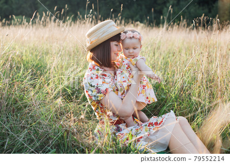 Mother, sitting on the grass in the field outdoor and hugging her little cute baby daughter. Mother, sitting on the grass in the field outdoor and hugging her little cute baby daughter. 79552214