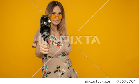 Cheerful young woman holding movie camera and posing against yellow background 79570704