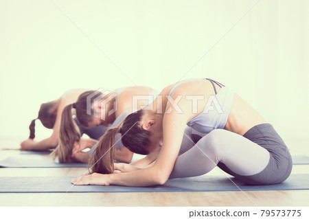 Group of young sporty attractive women in yoga studio, practicing yoga lesson with instructor, sitting on floor in forward bend yoga sana posture. Healthy active lifestyle, working out indoors in gym 79573775