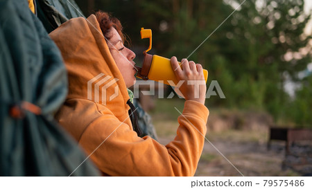 Caucasian woman drinking hot tea from a thermos while resting in a tourist tent. Caucasian woman drinking hot tea from a thermos while resting in a tourist tent. 79575486