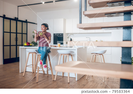 A dark-skinned woman sitting in the kitchen and eating salad A dark-skinned woman sitting in the kitchen and eating salad 79583364