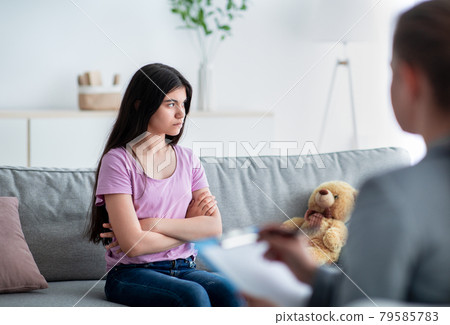 Unhappy Indian teen girl having session with psychologist at clinic, sitting with crossed arms, thinking about problems Unhappy Indian teen girl having session with psychologist at clinic, sitting with crossed arms, thinking about problems 79585783