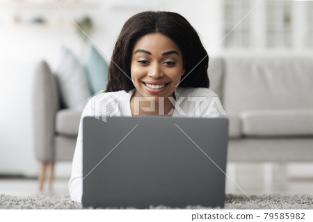 Remote job opportunities. Cheerful african american woman working on laptop at home, lying on floor in living room 79585982