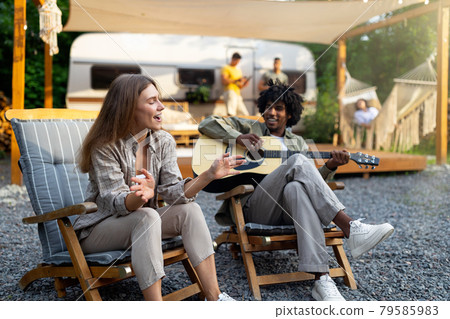 Black guy playing guitar, his female friend singing, sitting in lounge chairs near motorhome, having fun camping trip Black guy playing guitar, his female friend singing, sitting in lounge chairs near motorhome, having fun camping trip 79585983