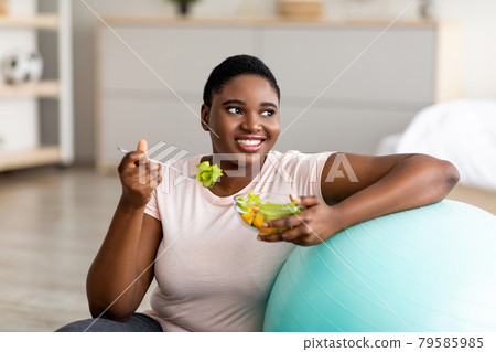 Sports and healthy nutrition for weight loss. Curvy black lady leaning on fitness ball, eating vegetable salad at home Sports and healthy nutrition for weight loss. Curvy black lady leaning on fitness ball, eating vegetable salad at home 79585985