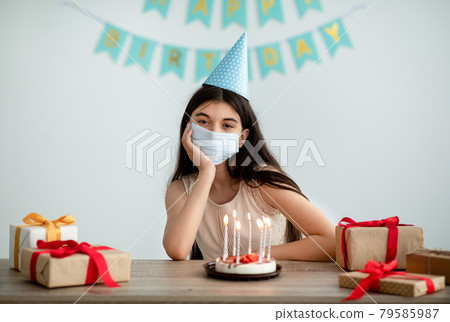 Unhappy Indian teenage girl in face mask and festive hat having dull birthday alone at home during covid isolation Unhappy Indian teenage girl in face mask and festive hat having dull birthday alone at home during covid isolation 79585987