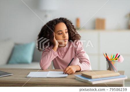 Thoughtful black teen girl writing in notepad, doing homework and looking away, sitting at table with books 79586239