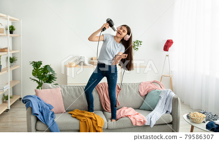 Indian teen girl wearing headphones, using blowdryer as microphone, singing song and dancing on sofa at home 79586307