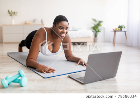 Plump black lady exercising at home, following personal trainer's online instructions on laptop, standing in plank pose Plump black lady exercising at home, following personal trainer's online instructions on laptop, standing in plank pose 79586334