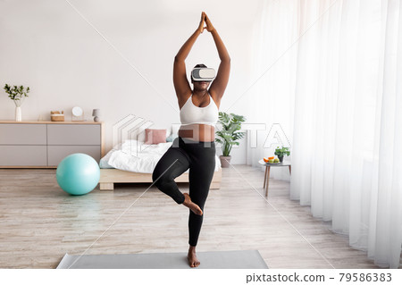 Overweight black lady wearing VR headset, practicing yoga in augmented reality at home Overweight black lady wearing VR headset, practicing yoga in augmented reality at home 79586383
