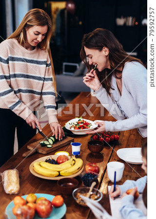 Two young women are preparing lunch, one is cutting vegetables for a salad, the other is tasting and the girl next to her is doing her homework. Two young women are preparing lunch, one is cutting vegetables for a salad, the other is tasting and the girl next to her is doing her homework. 79592757