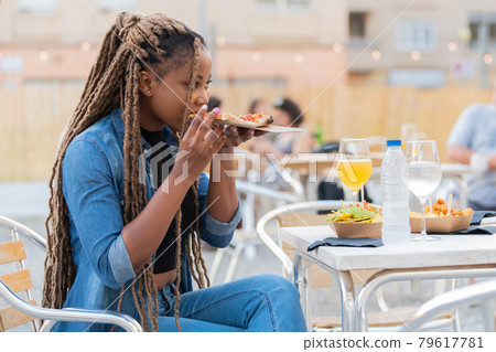 Cute afro american woman enjoying a pizza slice and rinks at an outdoors terrace 79617781