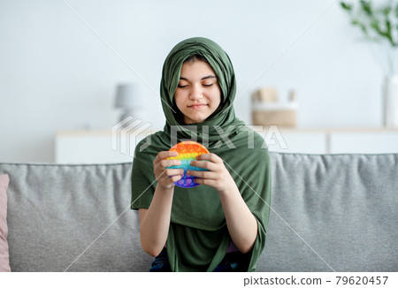 Portrait of lovely Indian teen girl in hijab playing with bright POP IT toy, enjoying fun antistress game at home Portrait of lovely Indian teen girl in hijab playing with bright POP IT toy, enjoying fun antistress game at home 79620457