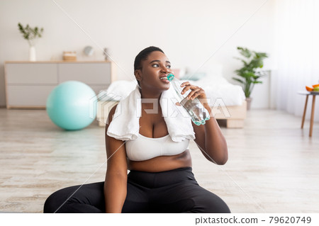 Wellness and weight loss. Plus size black lady drinking fresh clear water from bottle after sports workout at home 79620749