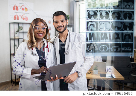 Afro-american lady and arabian man, standing at bright office, working on laptop and smiling Afro-american lady and arabian man, standing at bright office, working on laptop and smiling 79636495