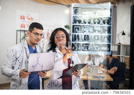 Multiracial doctors examining tomography of patient, standing behind glass wall at office Multiracial doctors examining tomography of patient, standing behind glass wall at office 79636501