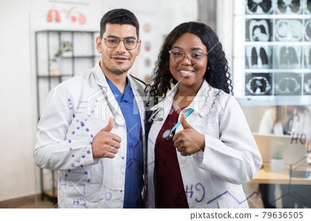 Confident young doctors, Afro-American woman and Indian man, posing at bright office with thumbs up 79636505