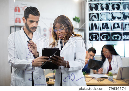 Two multiethnic male and female doctors standing behind glass wall and working on tablet pc Two multiethnic male and female doctors standing behind glass wall and working on tablet pc 79636574