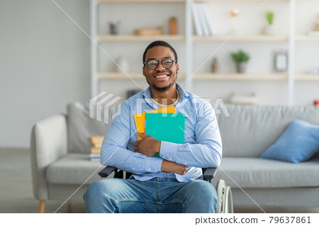 Positive disabled black in wheelchair holding notebooks and smiling at camera at home 79637861