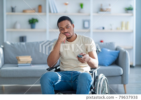Disabled black man in wheelchair using remote control, watching dull movie on TV, cannot find interesting program 79637938