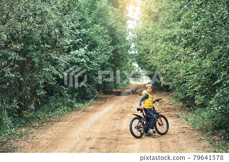 Boy in vest with bicycle looks at camera on rural road Boy in vest with bicycle looks at camera on rural road 79641578