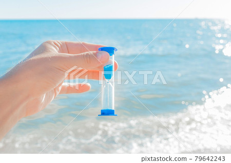 The hand holds the hourglass against the blue bright sea and the sky with a horizon line, close-up 79642243