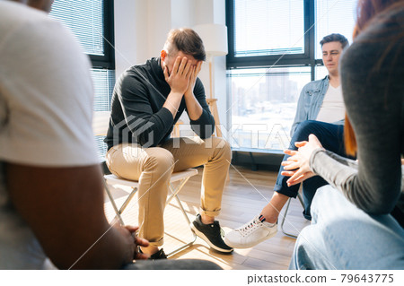 Sad depressed male talking with compassionate patients and psychotherapist sitting in circle during group interpersonal therapy session. Sad depressed male talking with compassionate patients and psychotherapist sitting in circle during group interpersonal therapy session. 79643775