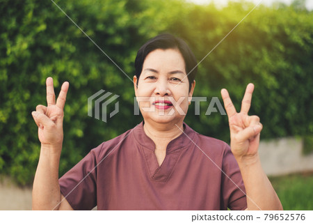 Mature asian woman standing and showing 2 fingers at public park,Happy and smiling,Senior care insurance concept 79652576