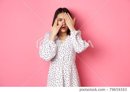 Disappointed asian woman peeking through fingers, open eyes and frowning displeased, staring with disdain at camera, standing over pink background Disappointed asian woman peeking through fingers, open eyes and frowning displeased, staring with disdain at camera, standing over pink background 79659383