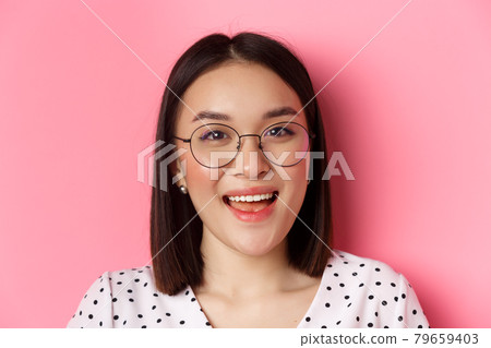 Beauty and lifestyle concept. Close-up of cute asian female model wearing trendy glasses, smiling happy at camera, standing on pink background Beauty and lifestyle concept. Close-up of cute asian female model wearing trendy glasses, smiling happy at camera, standing on pink background 79659403