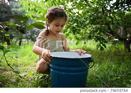 Cute baby girl sitting in the garden and peeking into a metal bucket 79659784