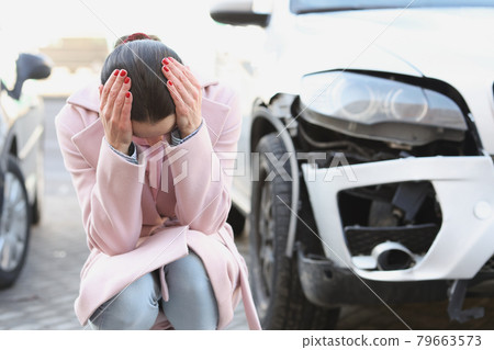 Upset young woman sits with head bowed next to wrecked car Upset young woman sits with head bowed next to wrecked car 79663573