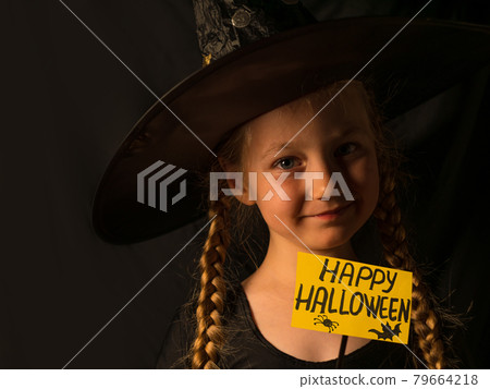 Little pretty girl witch in black wizard hat holds greeting Happy Halloween card smiling. Autumn kids party celebration. 79664218