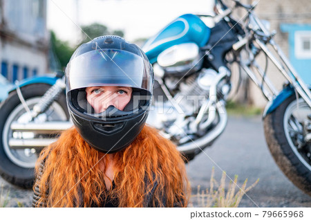 Red-haired curly woman in a helmet near a motorcycle. 79665968