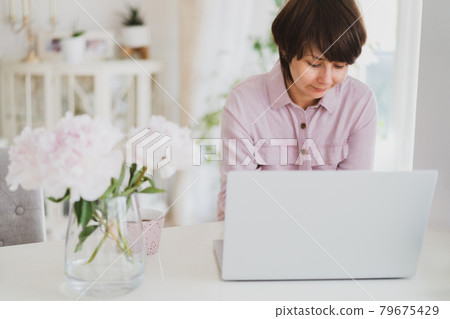 Mixed race woman working at home using a laptop. Cozy workspace with fresh pink peonies flowers and a cup of tea on the white table with light interior background. Remote work at home. Copy space Mixed race woman working at home using a laptop. Cozy workspace with fresh pink peonies flowers and a cup of tea on the white table with light interior background. Remote work at home. Copy space 79675429