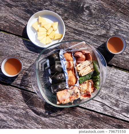 Picnic table made of rough old wood with cups of tea, a bowl of pineapple and a bowl of sushi, sausages, egg and asparagus Picnic table made of rough old wood with cups of tea, a bowl of pineapple and a bowl of sushi, sausages, egg and asparagus 79675550