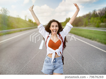 Excited young woman walking along highway, lifting hands up and shouting, feeling happy and free on summer journey Excited young woman walking along highway, lifting hands up and shouting, feeling happy and free on summer journey 79676269
