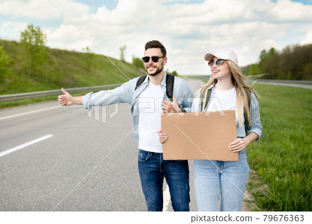 Happy millennial couple standing along highway with empty sign, stopping car, having road trip, mockup for design 79676363