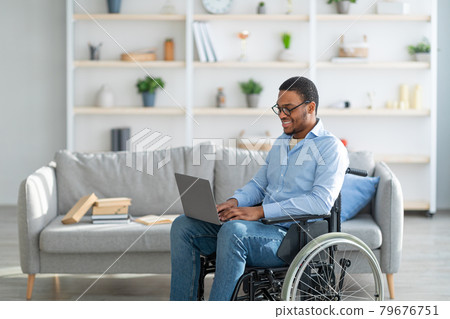 Cheerful handicapped black guy in wheelchair using laptop for work or studies, having remote job at home, copy space 79676751