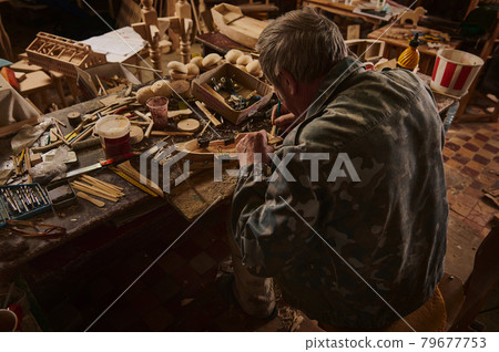 Rear view of an old craftsman making a wooden model of sailship in his home workshop. Carpenter at work Rear view of an old craftsman making a wooden model of sailship in his home workshop. Carpenter at work 79677753
