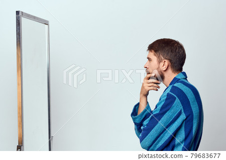 a man in a blue robe examines himself in a mirror in a bright room cropped view a man in a blue robe examines himself in a mirror in a bright room cropped view 79683677