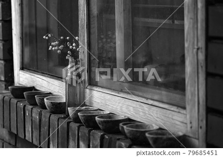 A bouquet of lilies of the valley in a glass bottle stands on a brick windowsill on the street. There are many small earthenware cups next to the bottle. Wooden window. Black and white photography 79685451