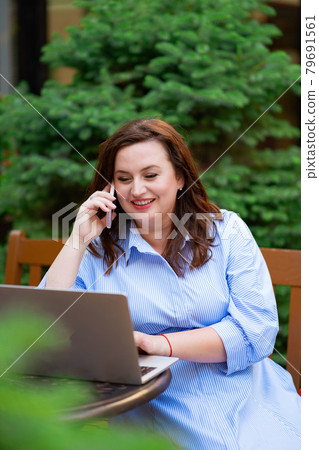 woman sits on an outdoor cafe and works on a laptop and talks on the phone 79691561