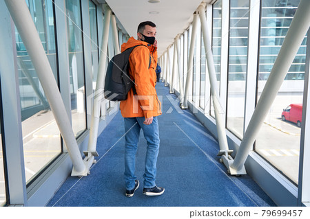 Guy tourist with a backpack and in a medical mask is walking along the airbridge from the plane to the airport terminal Guy tourist with a backpack and in a medical mask is walking along the airbridge from the plane to the airport terminal 79699457