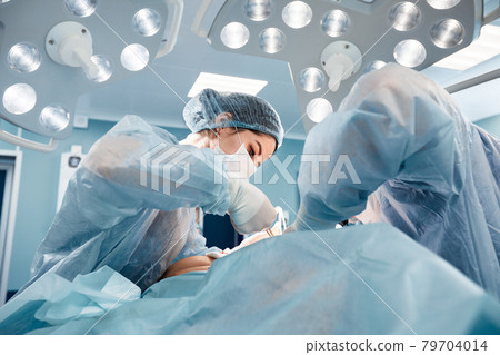 Surgeons men and women in one team during an operation over the operating table, doctors in modern equipment perform an oncological operation Surgeons men and women in one team during an operation over the operating table, doctors in modern equipment perform an oncological operation 79704014