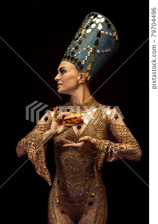 Young beautiful woman in image of Nefertiti in art performance tasting modern fast food isolated on dark background. Retro style, comparison of eras, humor concept. 79704496