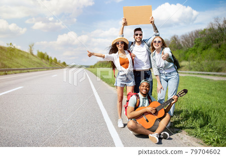 Cheerful diverse friends with backpacks and guitar hitchhiking on highway, catching free ride, having summer adventure Cheerful diverse friends with backpacks and guitar hitchhiking on highway, catching free ride, having summer adventure 79704602