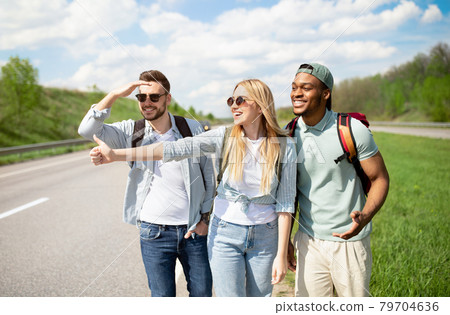 Cheerful diverse friends trying to get free ride, hitchhiking on road, showing thumb up outdoors Cheerful diverse friends trying to get free ride, hitchhiking on road, showing thumb up outdoors 79704636